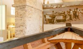 Se aprecia dentro de un museo de piedra, la parte delantera de una barca de madera de pescadores y un cuadro colgado en la pared que es una fotografía de varios pescadores sacando una barca a la orilla